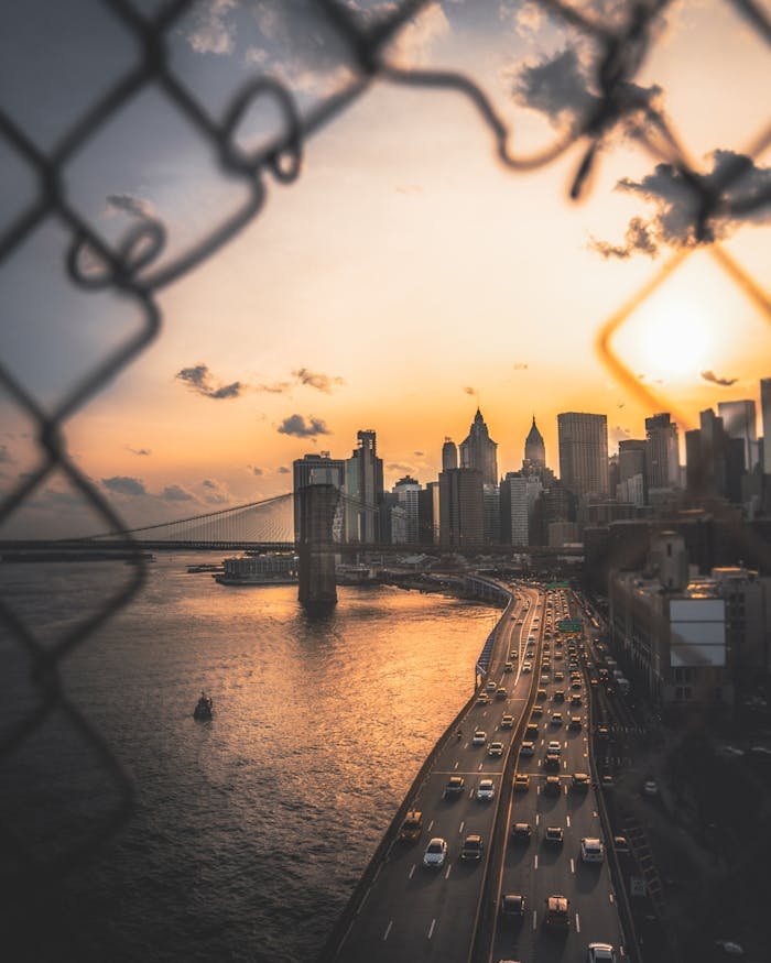 Captivating view of New York City skyline with Brooklyn Bridge during sunset, framed artistically by chain-link fence.